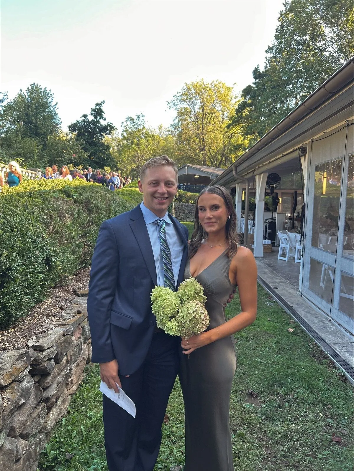 Couple dressed for a wedding outdoors, she holds white hydrangeas