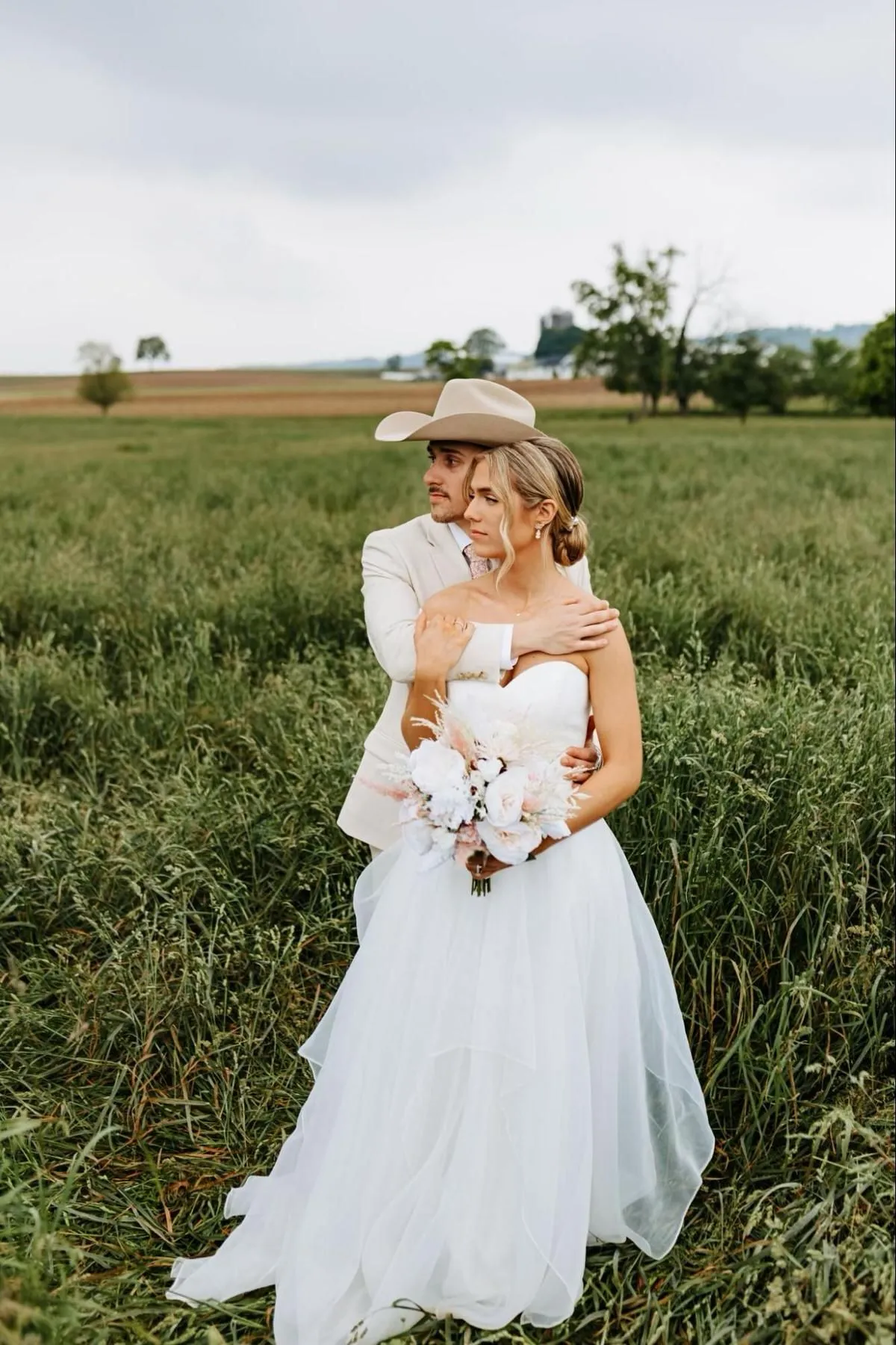 Groom in a cowboy hat hugging the bride from behind in a grassy field