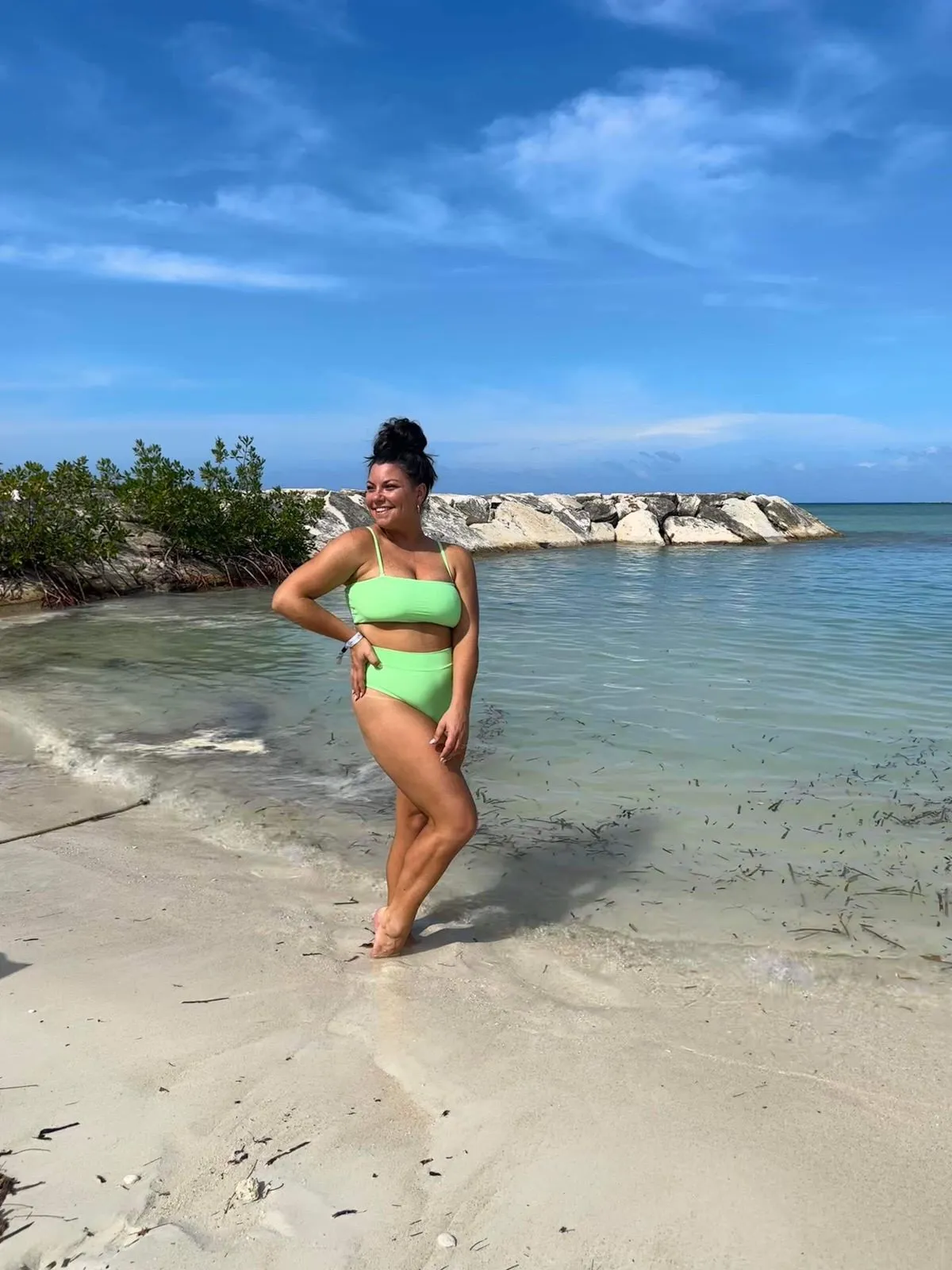 Woman in a green bikini standing on a beach by clear blue water