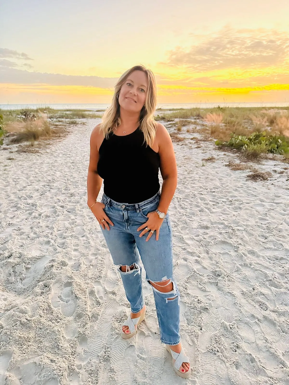 Woman in a black top and ripped jeans on a sandy beach path at sunset