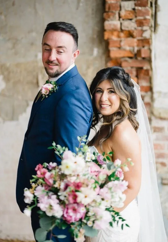 Groom in a navy suit with the bride holding a pink bouquet by a brick wall