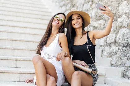 Two friends in summer outfits smiling and taking a selfie on stone steps